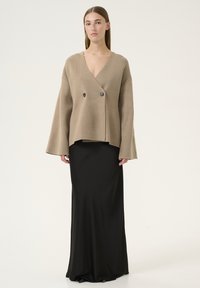 Cardigan overdimensionato beige in lana con doppio bottone, abbinato a una gonna nera maxi aderente, caratterizzata da una texture liscia e maniche lunghe.