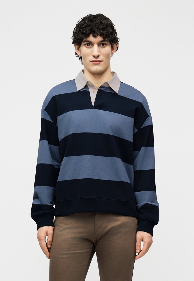 Jeune homme aux cheveux bouclés portant un maillot de rugby rayé bleu et noir avec un col beige et un pantalon marron, faisant face à l'avant.