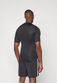 Svart kortermet treningstrøye med en tett passform, laget av strukturert materiale. Brukt med svarte shorts med hvite sidestriper.