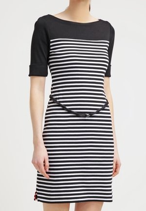 Femme portant une robe à rayures horizontales noires et blanches avec des manches courtes noires et une fine ceinture noire à la taille.