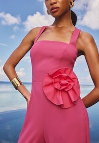 Roze jumpsuit met een vierkante halslijn, brede bandjes en een opvallend ruches bloemdetail op de heup; gecombineerd met een gouden manchetarmband.