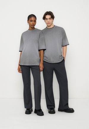 Δύο μοντέλα στέκονται δίπλα δίπλα φορώντας ταιριαστά oversized γκρι μπλουζάκια, σκούρα γκρι παντελόνια και μαύρα χοντρά παπούτσια πάνω σε απλό λευκό φόντο.