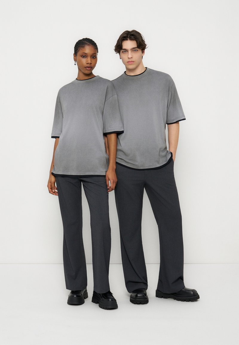 Δύο μοντέλα στέκονται δίπλα δίπλα φορώντας ταιριαστά oversized γκρι μπλουζάκια, σκούρα γκρι παντελόνια και μαύρα χοντρά παπούτσια πάνω σε απλό λευκό φόντο.