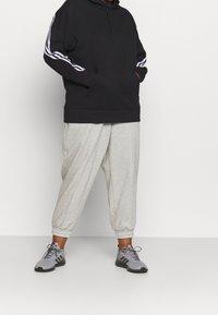 Personne portant un sweat à capuche noir avec des bandes blanches sur les manches, un pantalon de survêtement gris clair et des baskets grises avec des semelles noires, debout devant un fond blanc.