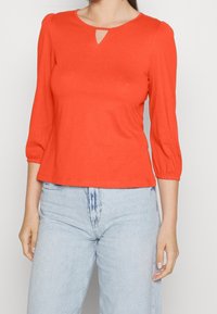 Red, fitted top with a keyhole neckline and three-quarter puff sleeves, paired with light blue denim jeans. Soft cotton material.