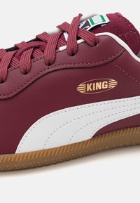 Burgunderfärgade lädersneakers med vit mockastripe och sula av gummigas. Har kastanjefärgade snören och en guldfärgad "KING"-logotyp på sidan.