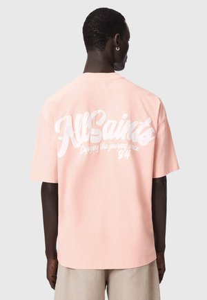 Mann trägt ein hellrosa oversized T-Shirt mit weißem Text „AllSaints Enjoying the journey since 94“ auf dem Rücken, beige Shorts.
