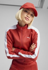 Röd sportjacka med vita ränder, dragkedja, hög krage och en liten logotyp. Matchande röd keps kompletterar outfiten. Slät textur.