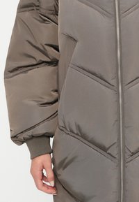 Chaqueta acolchada en un tono gris-marrón apagado, con cierre de cremallera en la parte frontal, costuras en forma de chevron y puños acanalados. El material parece brillante y aislante.