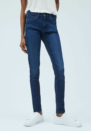 Osoba nosząca ciemnoniebieskie obcisłe dżinsy, białe trampki i białą bezrękawnik włożoną do spodni, stojąca na jednolitym tle.