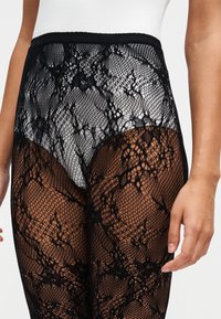 Gros plan d'une personne portant des collants noirs à motif de dentelle florale sur des sous-vêtements blancs, une main reposant sur le côté.