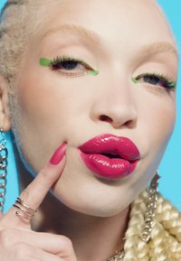 Gros plan d'une personne avec un eye-liner vert vif, des lèvres fuchsia brillantes, des cheveux blonds tressés, des boucles d'oreilles en chaîne argentée et un doigt manucuré rose touchant sa joue.