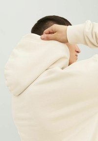 Hoodie w kolorze kremowym o teksturowanym materiale, z kapturem na sznurki, mankietami z ribbingu i luźnym krojem, pokazany z boku.