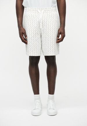 Modelo masculino con pantalones cortos blancos con estampado repetido "AX" en gris, combinado con calcetines blancos y zapatillas blancas, posando frente a un fondo blanco liso.