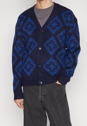 Cardigan navy con motivi a rombi blu, tessuto a maglia testurizzata, chiusura con bottoni e polsini a costine. Indossato sopra una t-shirt grigia.