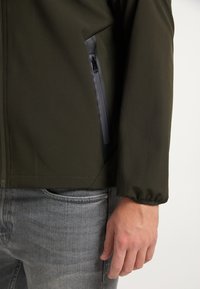 Olivgrön jacka med en slät textur, med en ficka med dragkedja i svart accent. Ärmen har en design med samlad manschett.