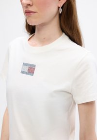 Hvid bomulds t-shirt med et lille firkantet logo med røde, blå og sorte detaljer, rund halsudskæring og korte ærmer.
