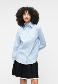 Camisa de botões de um azul claro com mangas longas, um único bolso no peito e botões escuros, combinada com uma saia pregueada preta com acabamentos brancos.
