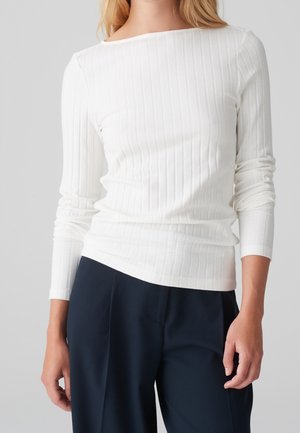 Femme portant un haut à manches longues blanc côtelé rentré dans un pantalon taille haute bleu marine, debout devant un fond uni.