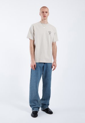 Giovane uomo con indosso una t-shirt beige con un motivo a cuore, jeans blu larghi, scarpe nere e collana e braccialetto d’argento, in piedi davanti a uno sfondo bianco.