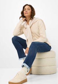 Cardigan beige a coste con bottoni sopra una sciarpa chiara, abbinato a jeans blu scuro e sneakers crema. Seduto su un pouf crema di texture.