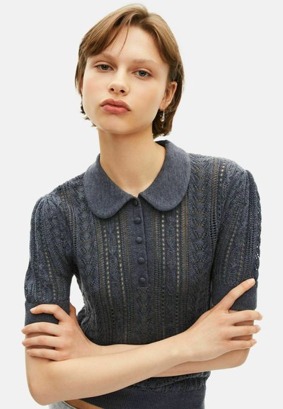 Młoda kobieta z krótkimi brązowymi włosami, nosząca ciemnoszary, krótko rękawowy sweter z kołnierzykiem w stylu Peter Pan, z założonymi na siebie rękami.