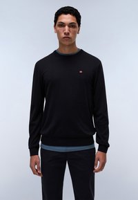 Jersey negro con cuello redondo, mangas largas y puños acanalados. Presenta un pequeño logo rojo y blanco en el pecho. Llevado sobre una camisa azul.