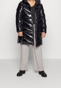 Schwarze, glänzende Daunenjacke mit Kapuze, ausgestattet mit Zickzack-Nähten und einem Reißverschluss. Getragen über einem grauen Oberteil und hell gestreiften Hosen.