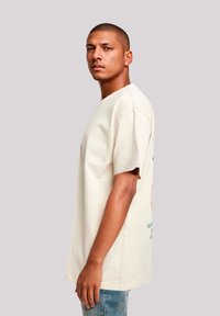 Crème korte mouwen t-shirt met ronde hals, met groene en oranje opdrukken op de achterkant, gedragen met lichtblauwe jeans.