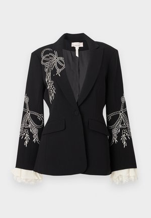 Kobieta ubrana w czarne spodnie i marynarkę na miarę z białym kwiatowym haftem, białą bluzkę wykończoną koronką oraz czarne szpilki stojąca na jednolitym tle.