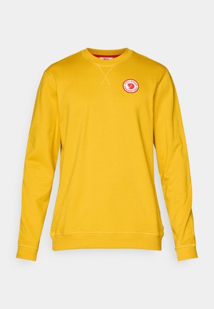 Gul sweatshirt lavet af blødt stof, med rund halsudskæring, lange ærmer, og et cirkulært logo-patch på brystet.