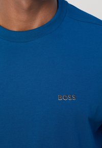 Blå bomullströja med ribbad rund hals och silverfärgad "BOSS"-logotyp på vänster bröst. Slät textur, figurnära design.