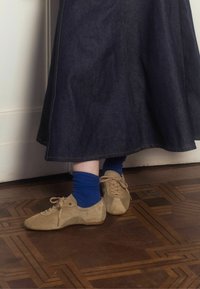 Scarpe in suede beige con punte arrotondate e lacci, indossate con calze a coste blu, abbinate a una gonna in denim scuro su un pavimento in legno con motivo.