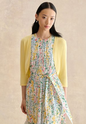 Mujer con cabello negro liso, que lleva pendientes de perlas, una chaqueta corta amarilla y un vestido floral atado en la cintura con una faja.