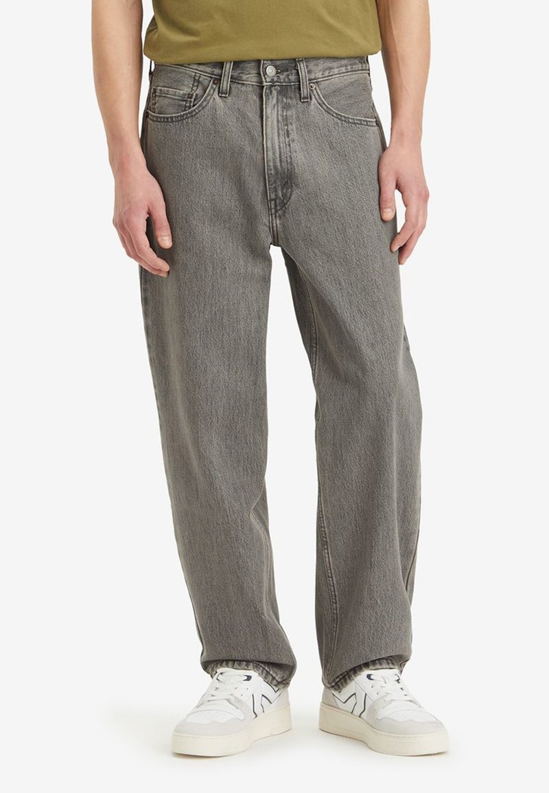 Vaqueros de mezclilla gris de corte holgado, con cinco bolsillos y cierre de botón en la parte delantera. Combinados con zapatillas blancas. Textura suave con un acabado desgastado.