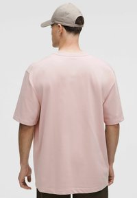 Hombre con una camiseta oversized de color rosa claro y una gorra de béisbol beige, visto desde atrás contra un fondo liso.