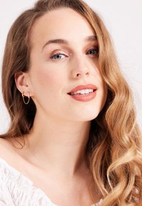 Boucles d'oreilles en or avec un accent en chaîne suspendue, accompagnées d'un haut en tissu léger. Le sujet a des cheveux bruns ondulés, arrivant aux épaules.