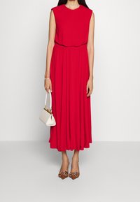 Czerwona, bezrękawna sukienka do kolan z marszczonym pasem. Zestawiona z brązowymi, spiczastymi szpilkami oraz małą białą torebką. Gładki materiał.