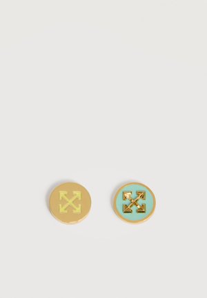 Dwa okrągłe metalowe przypinki z złotymi ikonami strzałek; jedna ma żółte tło, druga turkusowe tło.