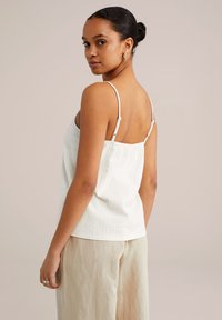 White textured camisole with thin adjustable straps, paired with loose beige trousers. The fabric has a subtle pattern and soft finish.