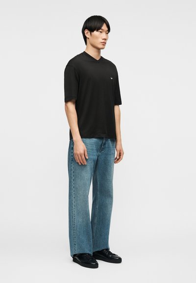 Νεαρός άνδρας όρθιος, φοράει μαύρο πουκάμισο με κοντά μανίκια, ανοιχτό μπλε τζιν με ίσια γραμμή και μαύρα αθλητικά παπούτσια, μπροστά σε απλό φόντο.