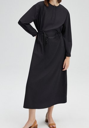 Kobieta w długiej czarnej sukience z długim rękawem i regulowanymi wiązaniami w talii, ubrana w jasnobrązowe sandały na słupku z odkrytymi palcami, pozująca na jednolitym tle.