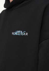 Hanorac negru cu glugă, cu text brodat albastru și alb pe piept, cu inscripția „FAVELA WORLD TOUR 2025”. Textură moale a țesăturii.