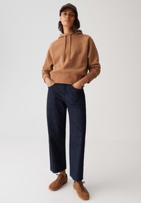 Persoon in een bruine hoodie, donkere blauwe wijde jeans, bruine sneakers en een donkere pet, staand met de handen in de zakken op een effen achtergrond.