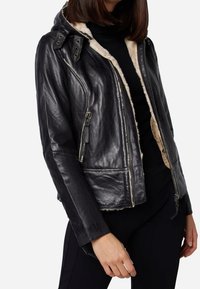 Giacca di pelle nera con cappuccio foderato in shearling, dotata di chiusura con zip, tasche laterali e dettagli di cuciture visibili.