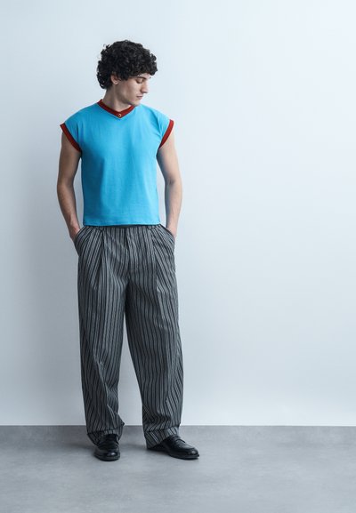 Jeune homme aux cheveux bouclés regardant vers le bas, portant un haut sans manches bleu avec des bordures rouges et un pantalon ample rayé, debout les mains dans les poches.