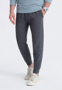 Pantalones de chándal grises con un ajuste ajustado, hechos de material suave, con detalles de parches, puños elásticos y bolsillos laterales. Combinados con zapatillas ligeras.