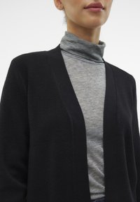 Cardigan nero con design aperto nella parte anteriore, indossato su un dolcevita grigio chiaro. Tessuto morbido, dettagli minimi e vestibilità rilassata.