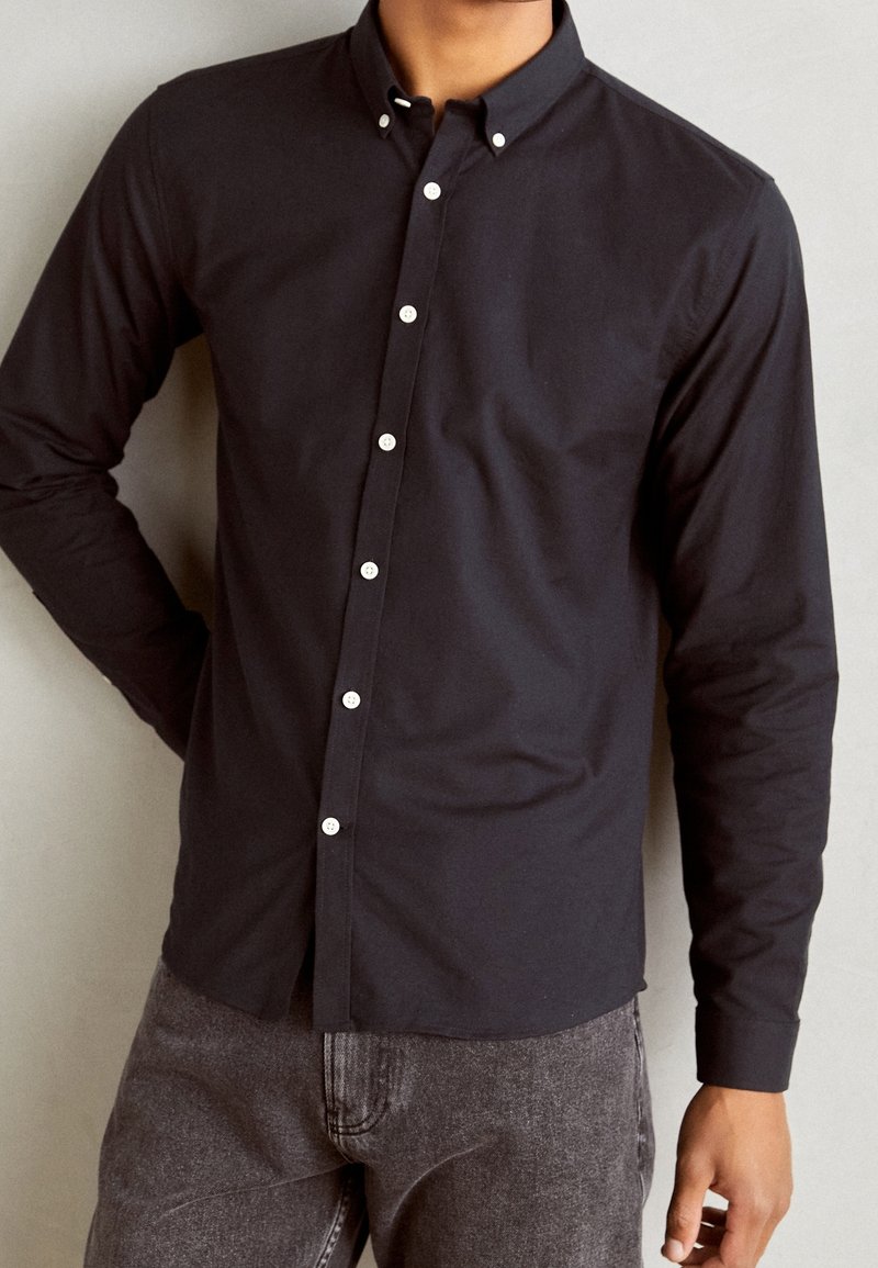 Homme portant une chemise noire à manches longues boutonnée, associée à un jean gris, debout devant un mur clair uni.