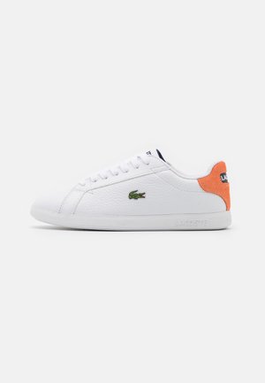 Baskets basses blanches avec un logo crocodile vert sur le côté et un empiècement en daim orange au talon, lacets blancs, et semelle plate blanche.